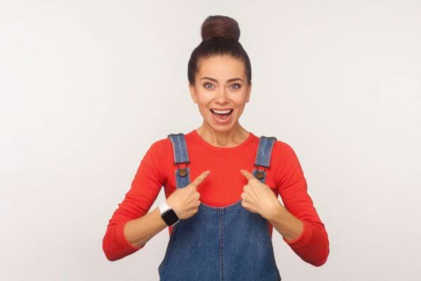 Une femme souriante en salopette et haut rouge se désigne avec enthousiasme du doigt.