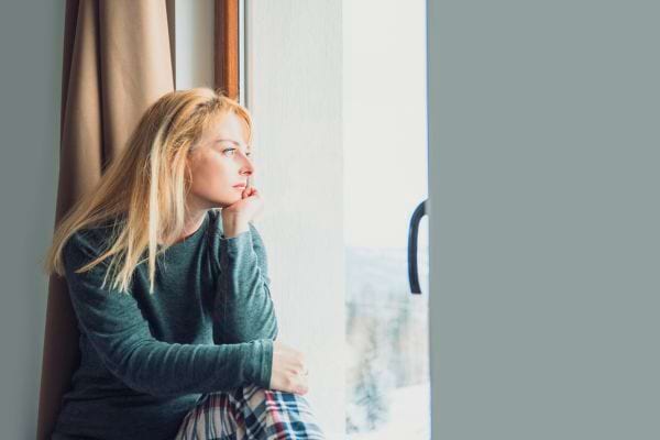 Une femme pensive regarde dehors assise près d’une fenêtre.