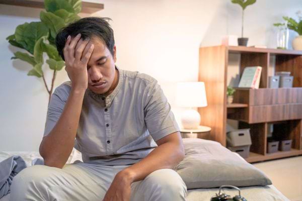 Un homme fatigué se tient la tête assis sur son lit.