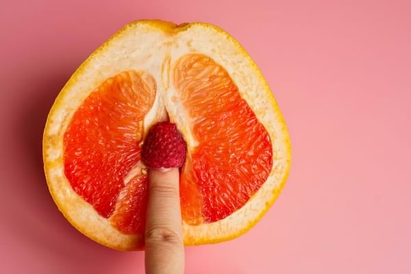 Un doigt touche une framboise placée dans un pamplemousse coupé, allusion suggestive.