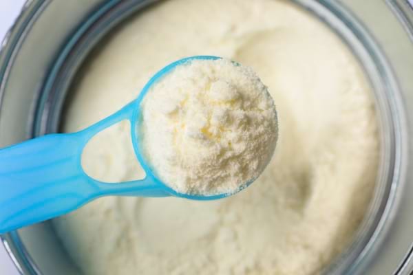 Une cuillère doseuse bleue contient du lait en poudre au-dessus d’une boîte métallique remplie.