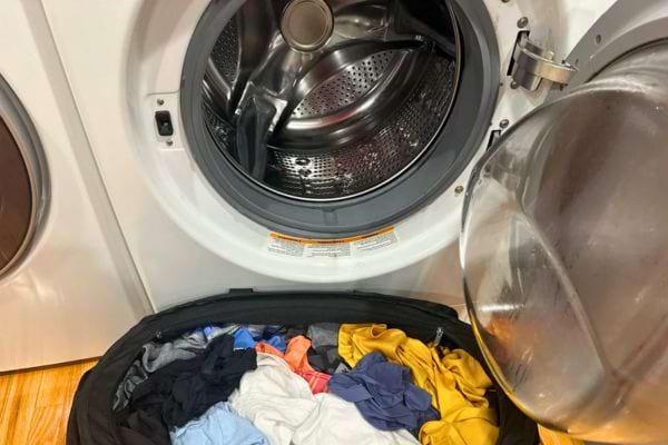 Un bac rempli de linge coloré est prêt devant une machine à laver ouverte.