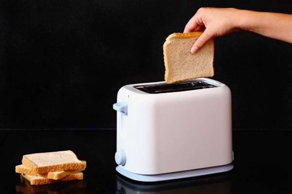 Une main insère une tranche de pain dans un grille-pain blanc.