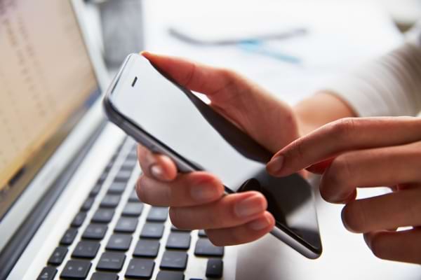 Une main tient un smartphone et tape dessus devant un ordinateur portable.