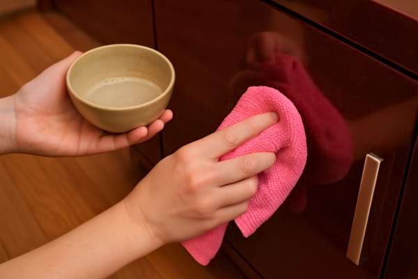 Une main nettoie un meuble en bois verni avec un chiffon rose et un petit bol de liquide.