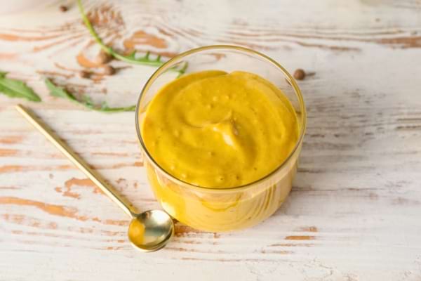 Un petit pot de moutarde jaune est posé sur une table en bois clair, accompagné d’une cuillère dorée.