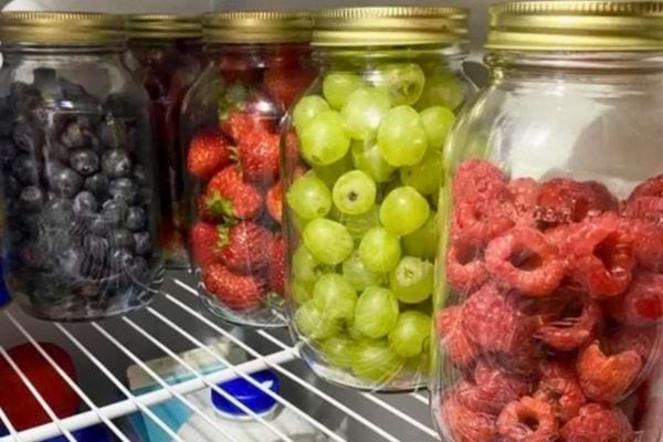 Des bocaux en verre remplis de raisins, fraises, myrtilles et framboises rangés dans un réfrigérateur.