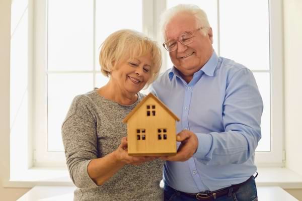 Couple de retraités souriant tenant une petite maison en bois.