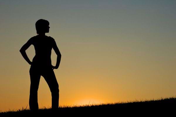 Une personne en silhouette se tient debout, mains sur les hanches, face à un coucher de soleil.