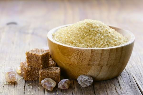 Un bol en bois clair contenant du sucre non raffiné accompagné de cubes bruns et morceaux de sucre brut.
