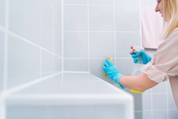 Une femme avec des gants bleus nettoie le carrelage blanc de la salle de bain avec un spray et une éponge.