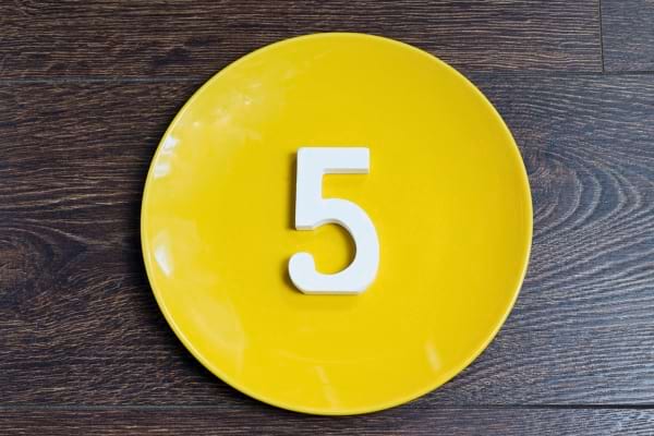 Un chiffre cinq blanc posé au centre d’une assiette jaune sur une table en bois sombre.
