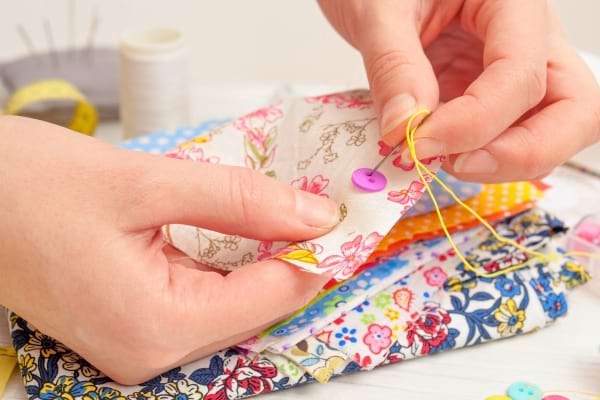 Des mains cousent un bouton rose sur un tissu fleuri coloré.