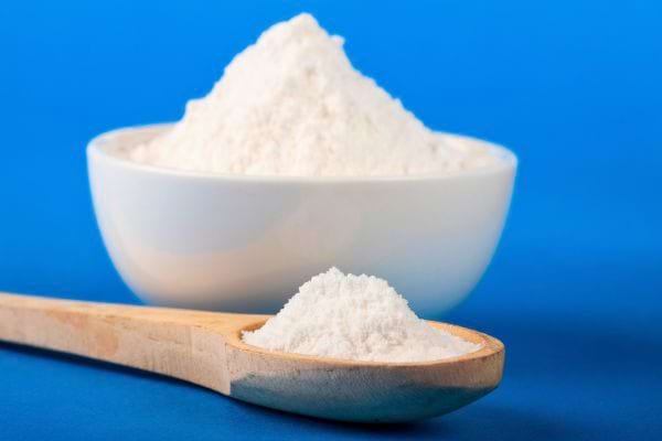 Une cuillère en bois remplie de poudre blanche devant un bol contenant de la même poudre.