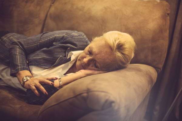 Une femme âgée s’assoupit sur un canapé, la tête appuyée sur sa main, vêtue d’une veste rayée.