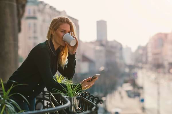 Une femme boit son café sur un balcon tout en tenant son téléphone.