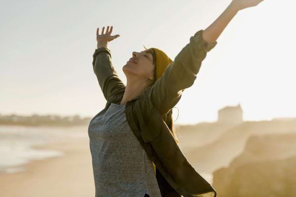 Une femme souriante lève les bras en signe de liberté au bord de mer.