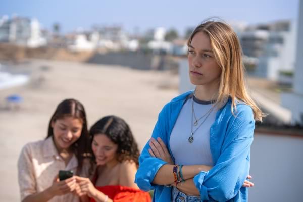 Une jeune femme croise les bras, à l’écart de deux amies qui rient ensemble.
