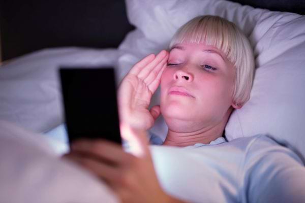 Une femme allongée dans son lit regarde son smartphone fatiguée.