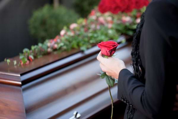 Une femme en noir tient une rose rouge près d’un cercueil fleuri.