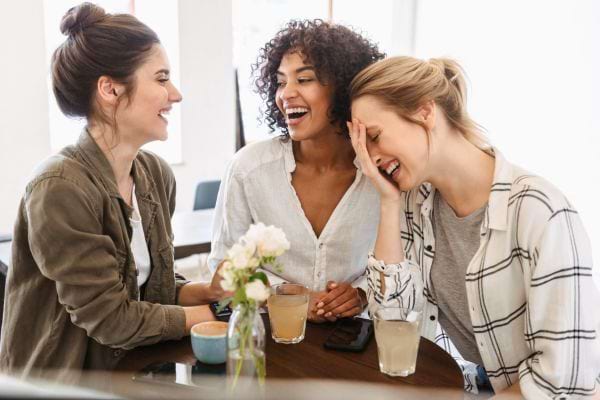 Trois amies rient ensemble autour de boissons dans un caf&eacute; chaleureux.