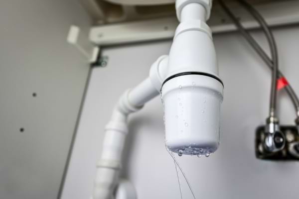 Un tuyau blanc sous un évier laisse couler quelques gouttes d’eau.