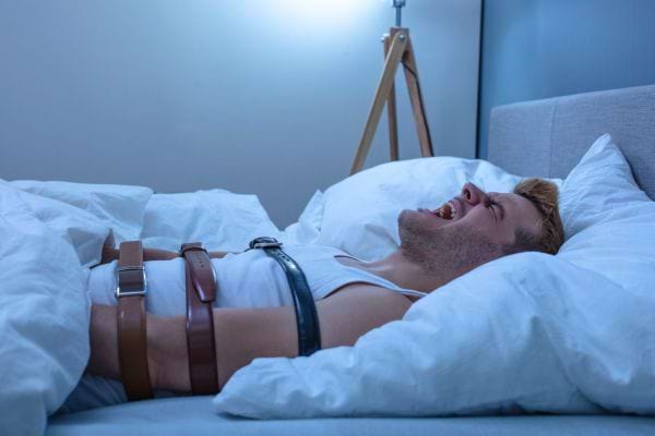 Un homme criant, immobilisé au lit avec des ceintures.