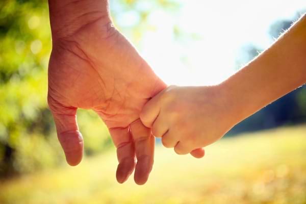 Une main adulte tient délicatement la main d’un enfant en extérieur.