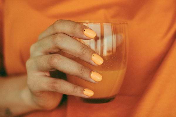 Main tenant un verre de jus orange assorti à un vernis coloré.