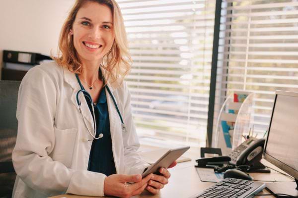 Une femme médecin sourit à la caméra, assise à son bureau avec une tablette en main.