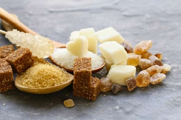 Un assortiment varié de sucres : cubes blancs, bruns, sucre roux, cristaux et morceaux sur une surface grise.