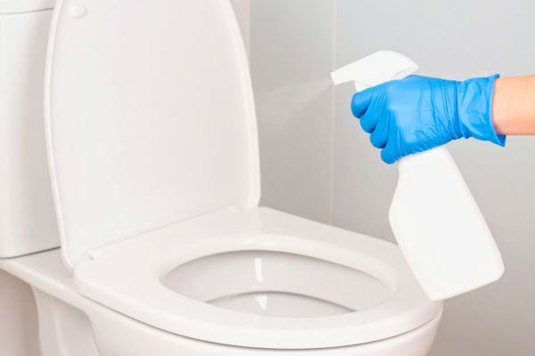 Une main avec gant bleu pulvérise un produit nettoyant sur une cuvette de toilette ouverte.