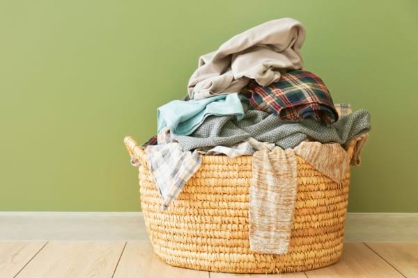 Un panier en osier déborde de vêtements sales colorés contre un mur vert.