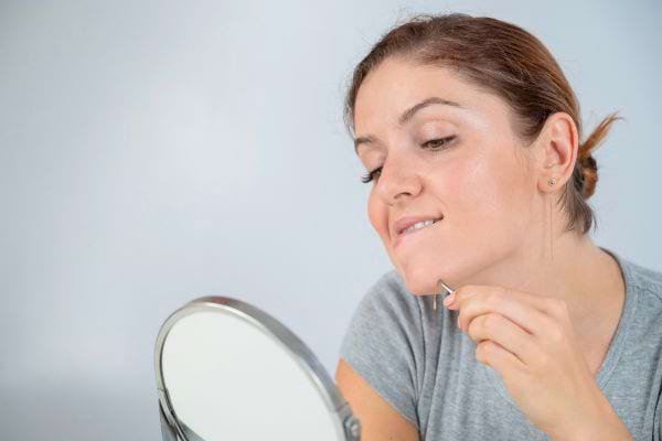 Une femme s’épile le menton devant un miroir avec une pince.