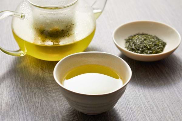 Une théière de thé vert accompagnée d’une tasse remplie et d’un petit bol de feuilles séchées.