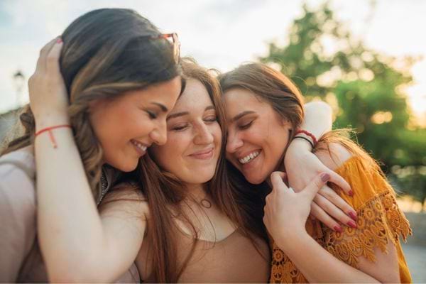 Trois amies souriantes s’enlacent en extérieur au coucher du soleil.
