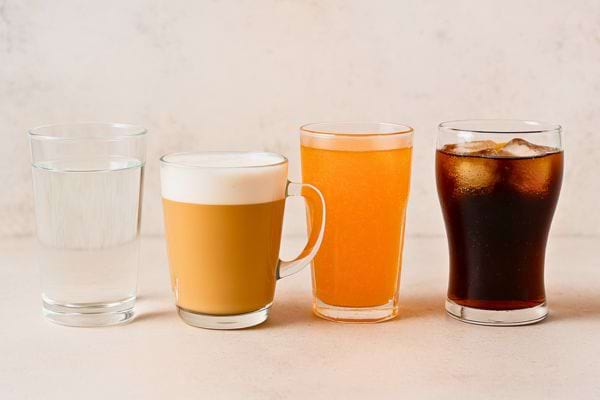 Quatre verres alignés contenant respectivement de l’eau, du café, du jus et du soda.