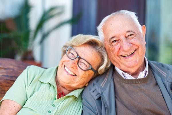 Un couple âgé souriant assis côte à côte à l’extérieur.