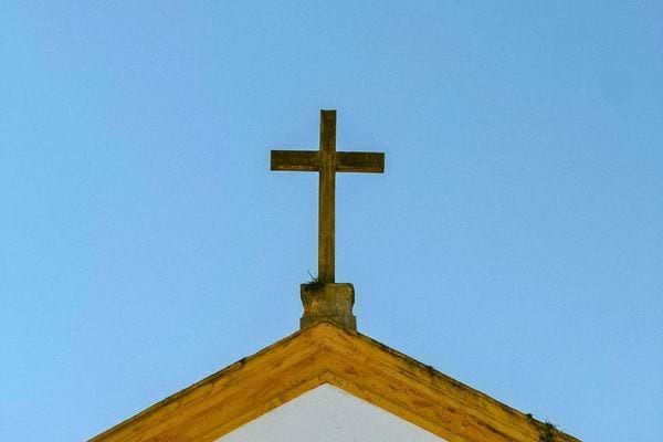 Une croix chrétienne en bois est posée au sommet d’un toit d’église sur fond de ciel bleu.
