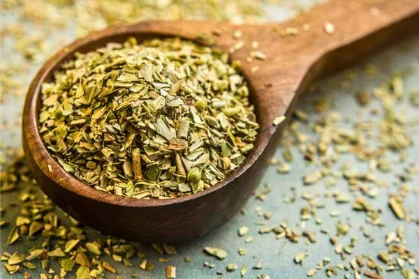 Une cuillère en bois remplie d’origan séché posée sur une surface bleue.