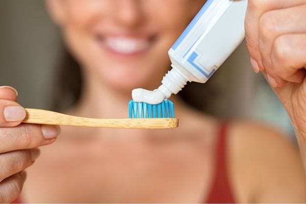 Femme souriante appliquant du dentifrice sur une brosse à dents en bambou, fond flou.