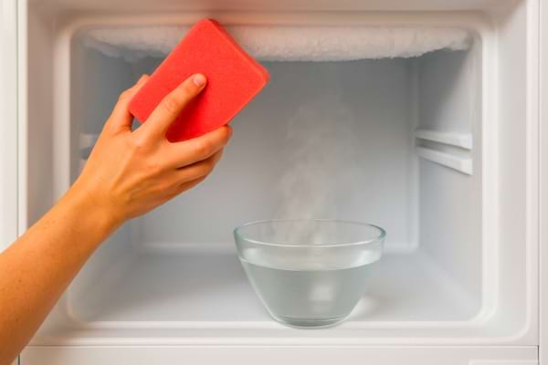 Une main dégivrant un congélateur, vapeur légère sortant d’un bol d’eau chaude.