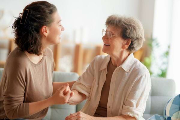 Une femme jeune parle et sourit à une femme âgée assise sur un canapé.