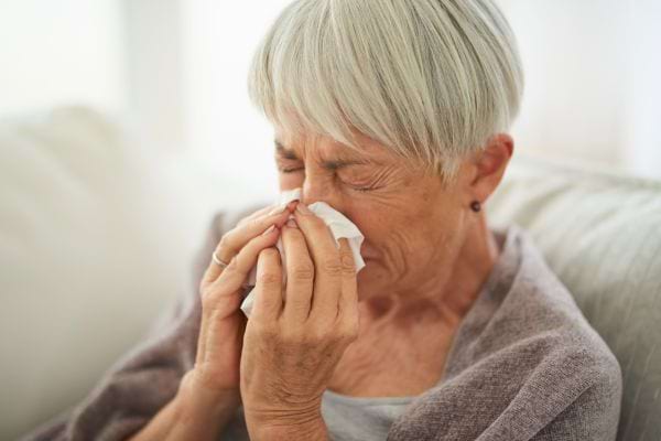 Une femme âgée se mouche avec un mouchoir en papier, les yeux fermés.