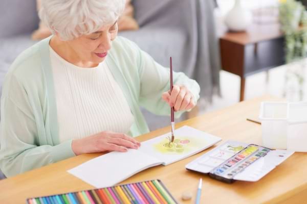 Une femme âgée souriante peint à l’aquarelle sur un carnet posé sur une table.