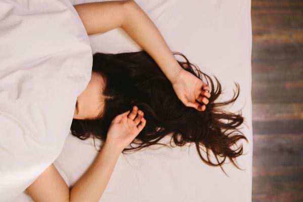 Une femme est cachée sous un drap blanc, laissant dépasser uniquement ses cheveux bruns étalés sur le lit.