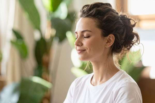 Une femme sourit les yeux fermés en respirant calmement dans un environnement lumineux.