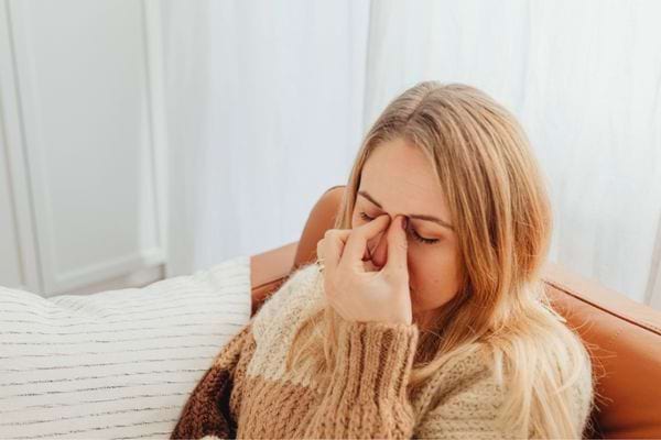 Femme fatiguée assise sur un canapé, se pinçant le nez à cause d’une migraine.
