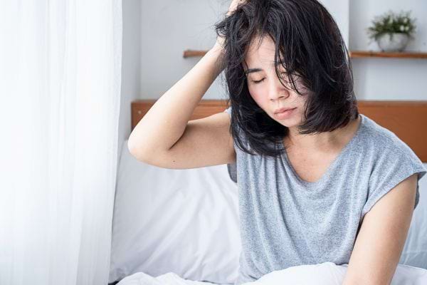 Une femme assise dans son lit, l’air fatigué, touche sa tête avec lassitude.