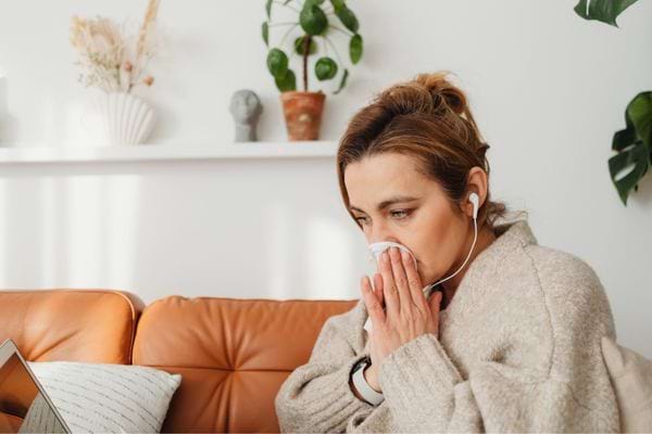 Femme en pull beige assise sur un canapé, se mouchant avec un mouchoir.